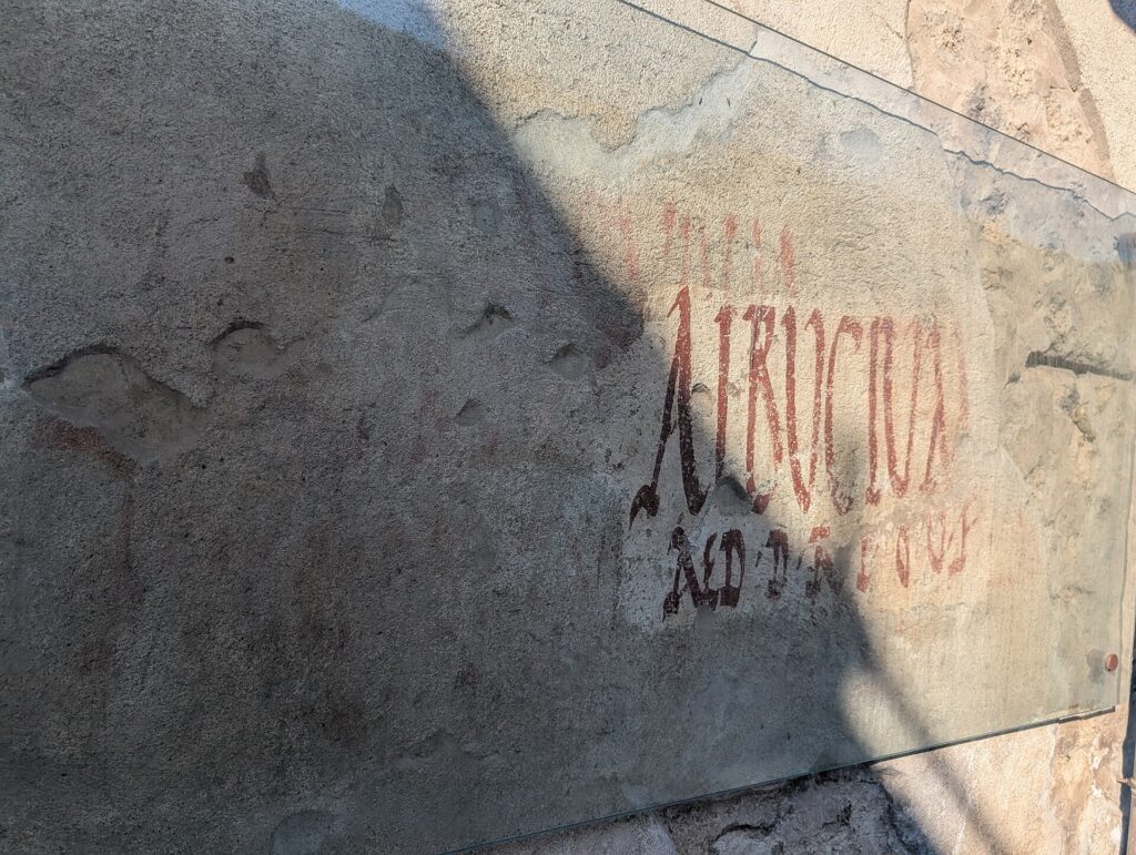 Regio V.3 Graffiti on the exterior wall. This thermopolium, excavated in 2019, features a typical serving counter, frescoed with a depiction of a Nereid riding a seahorse accompanied by darting dolphins, on its shorter side is an illustration of a figure moving between the different services offered by the thermopolium, a sort of enticing advertisement to attract the attention of passersby. On the outer side of the counter are depicted still lifes with depictions of animals, for example two mallard ducks shown upside down, probably slaughtered and sold here. The surface of the counter has circular recesses lined with walls of amphorae where food was stored, ready to be served. Analysis of remains found when these containers were emptied revealed the presence of pig, duck and fish bones - residues of the meals that were regularly prepared and sold here. Inside one of these containers a bottle for pouring and remains of fava beans used to whiten wine, were found.